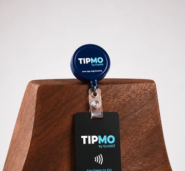 TIPMO Badge Reel — a branded retractable badge reel with an attached NFC tipping card reading tap here to tip, resting on a polished wood block